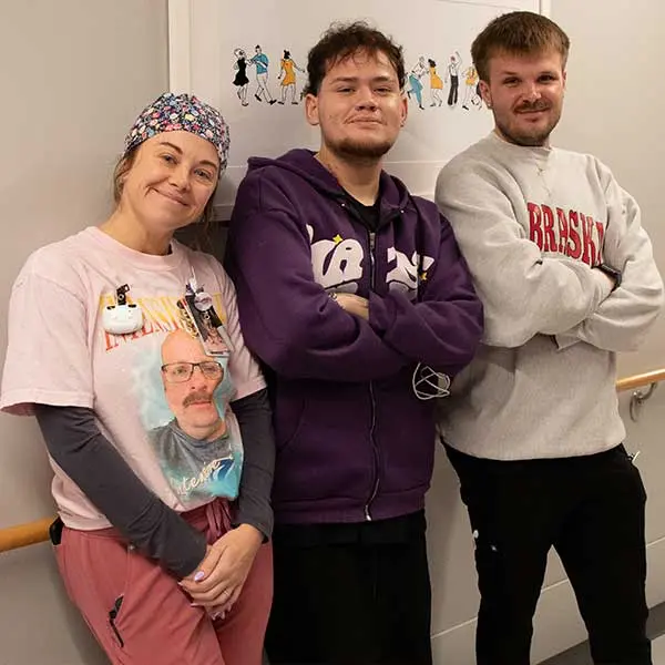 Three smiling people pose in a hospital hallway, including a woman in scrubs, Eduardo and a young men in casual sweatshirts.
