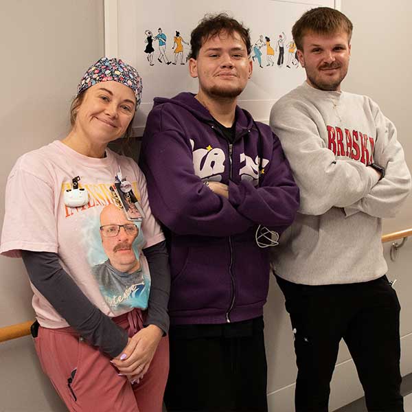 Three smiling people pose in a hospital hallway, including a woman in scrubs, Eduardo and a young men in casual sweatshirts.