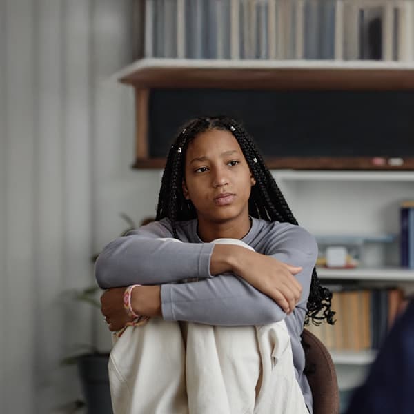 Person with braided hair in gray sweater sitting with arms wrapped around knees against bookshelf background.