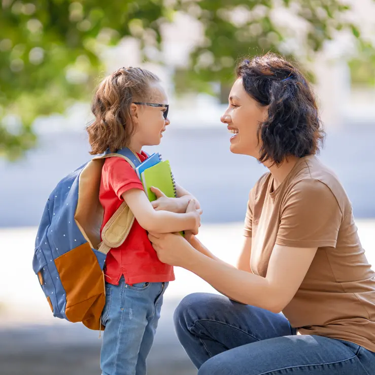 Parent talking to young child about school under a large shade tree, in a bright outdoor park setting.