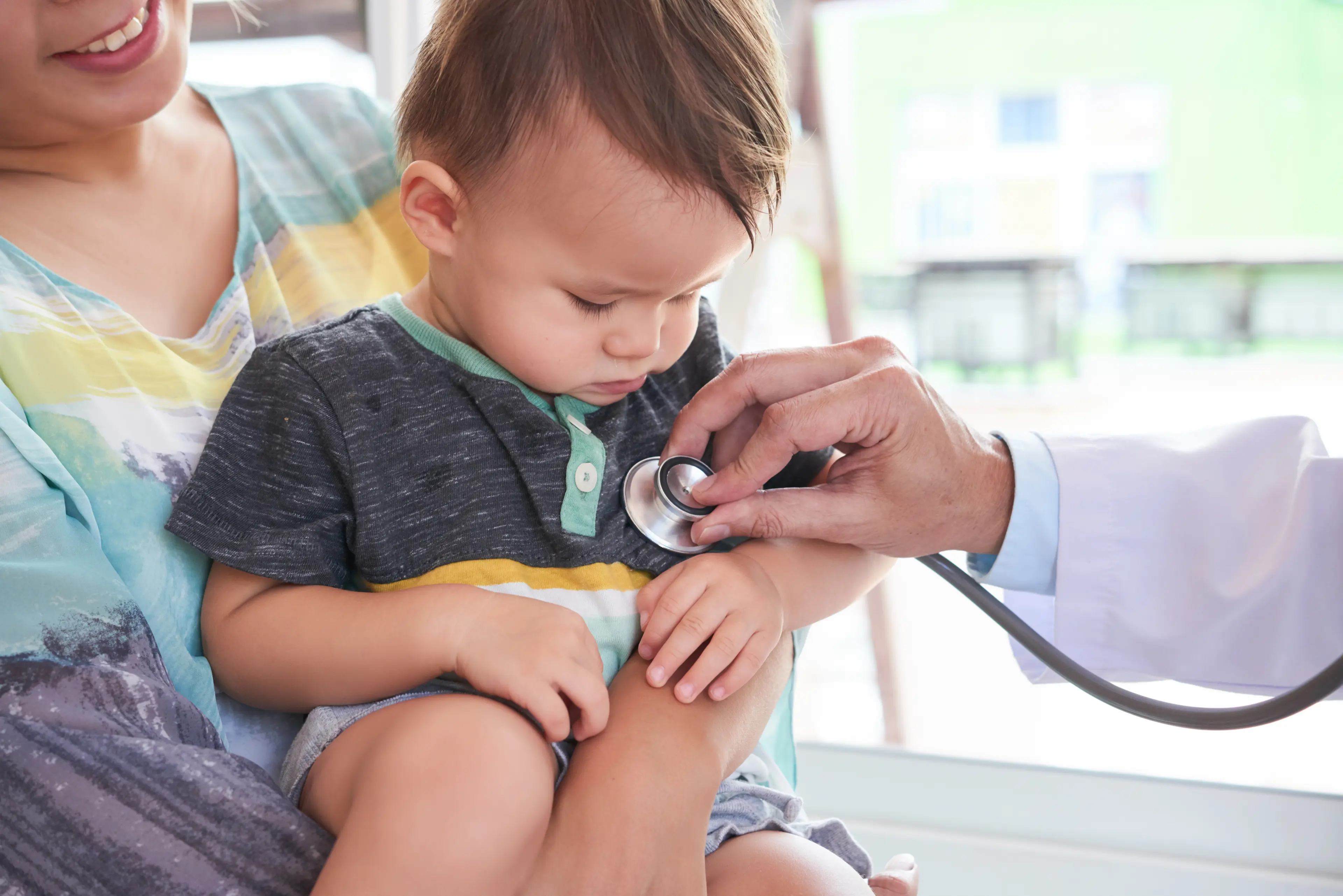 Cute toddler sitting on his mother's knees while doctor listening to his heart beat