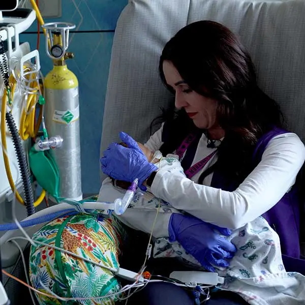 Sarah Freeland in blue gloves tenderly holds a small infant connected to medical tubes and monitors in a hospital setting.