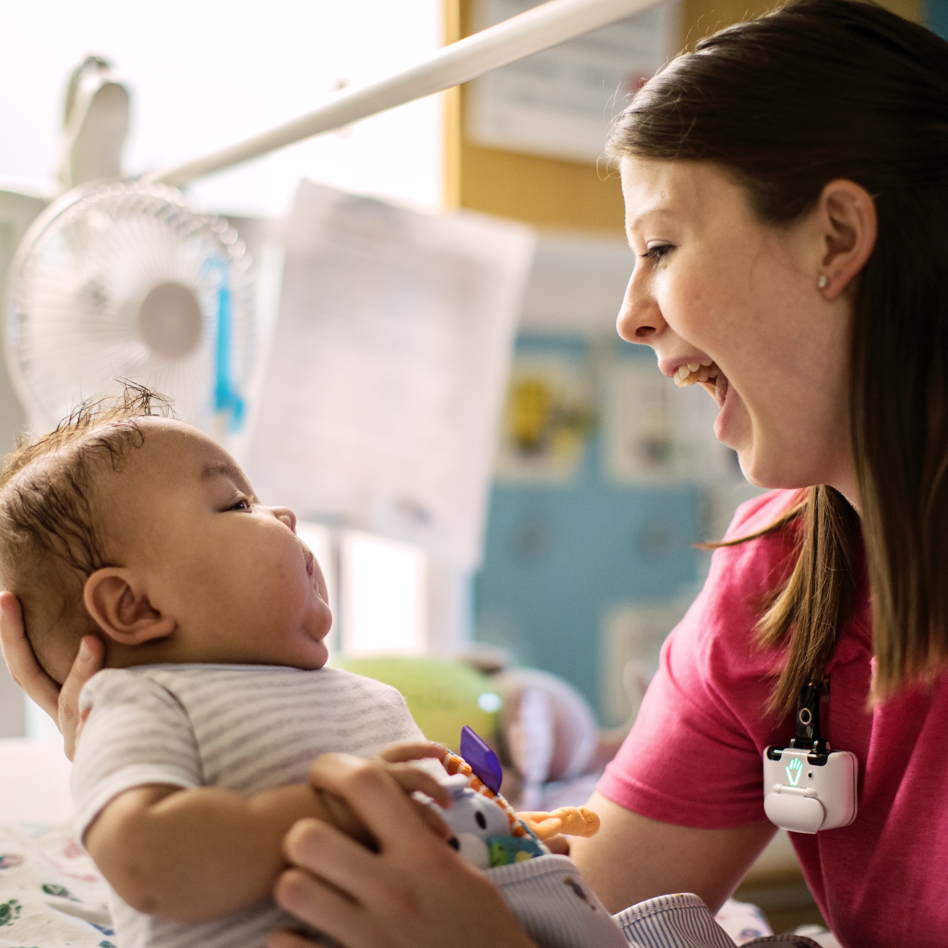 Woman in pink scrubs smiling at infant in hospital crib with colorful toys and mobile hanging above.