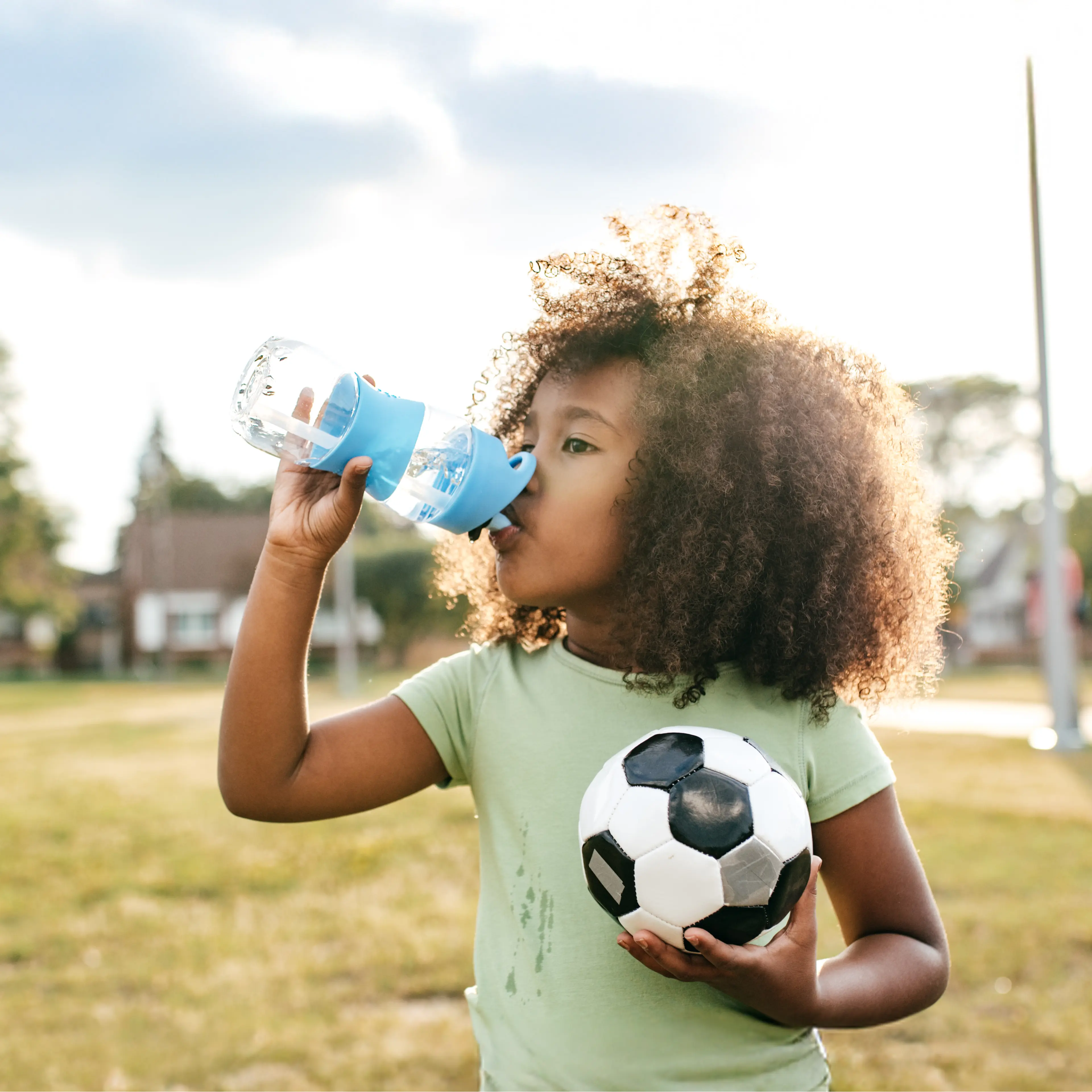 Child with curly hair drinking from blue water bottle while holding soccer ball in green shirt outdoors.