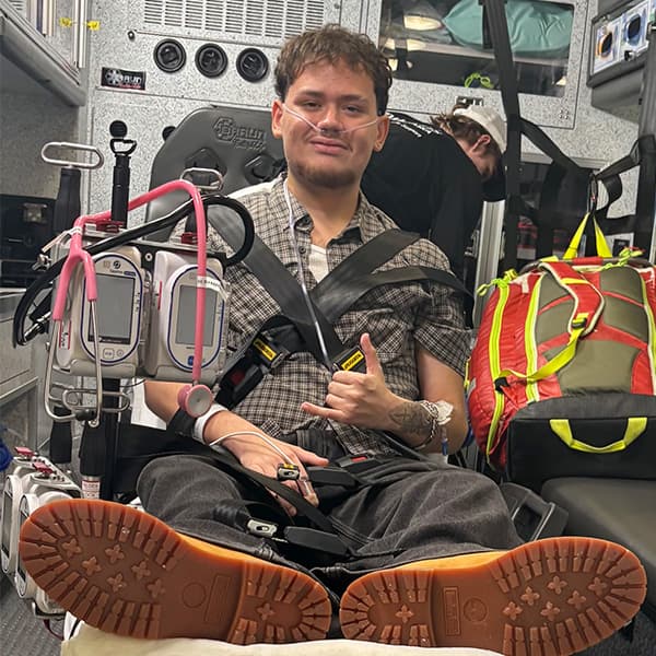 Eduardo with oxygen tube gives thumbs up while strapped in an ambulance with medical monitors nearby on his way to his high school graduation.