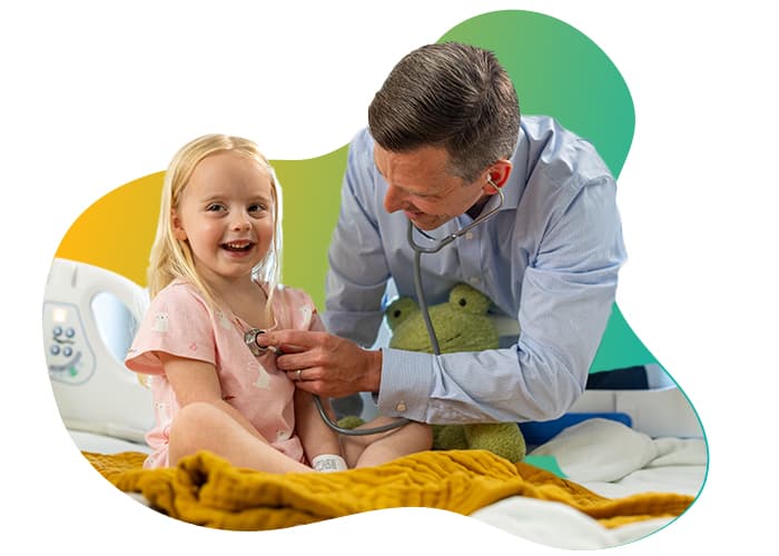 A smiling young girl in a pink dress sits on a hospital bed as a doctor uses a stethoscope during a checkup.