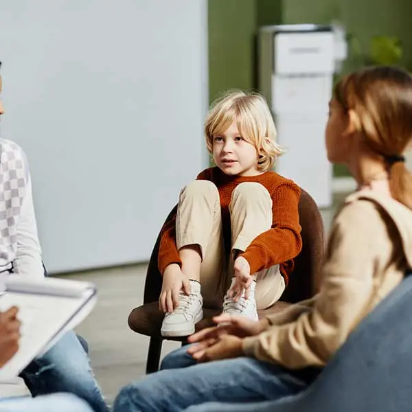 Young child in rust-colored sweater sitting in chair with knees pulled up, surrounded by people in a classroom setting.