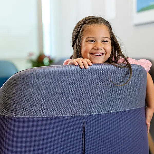 Young child with a bright smile peeking over the back of a blue chair in a bright indoor setting.