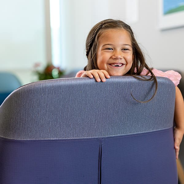 Young child with a bright smile peeking over the back of a blue chair in a bright indoor setting.