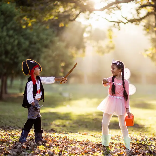 Children in pirate and fairy costumes playing in a sunlit forest clearing with autumn leaves on the ground.