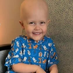 A young child with no hair smiling while wearing a blue patterned hospital gown.