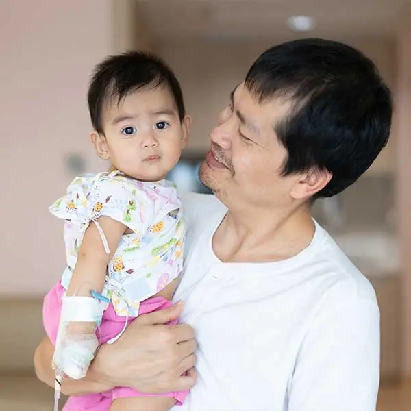 A person in a white shirt holding a baby wearing colorful hospital clothes with a medical IV attached to their arm.